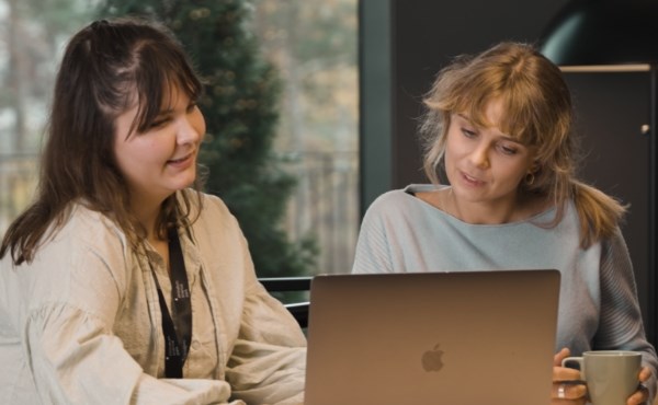 Två unga kvinnor tittar på samma datorskärm och pratar. Foto.