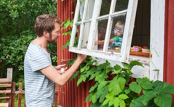 En pappa som pratar med sitt barn genom ett öppet fönster på en torpliknande byggnad