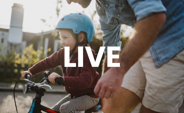 Barn med cykelhjälm, tränar på at cykla med hjälp av sin pappa. I bakgrunden syns hus och växter. Bildtext säger Live. Foto.