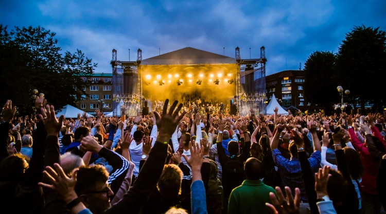 Konsert utomhus på torg i mörker. Publikhav i förgrunden framför scen lyfter sina händer mot skyn. Foto.