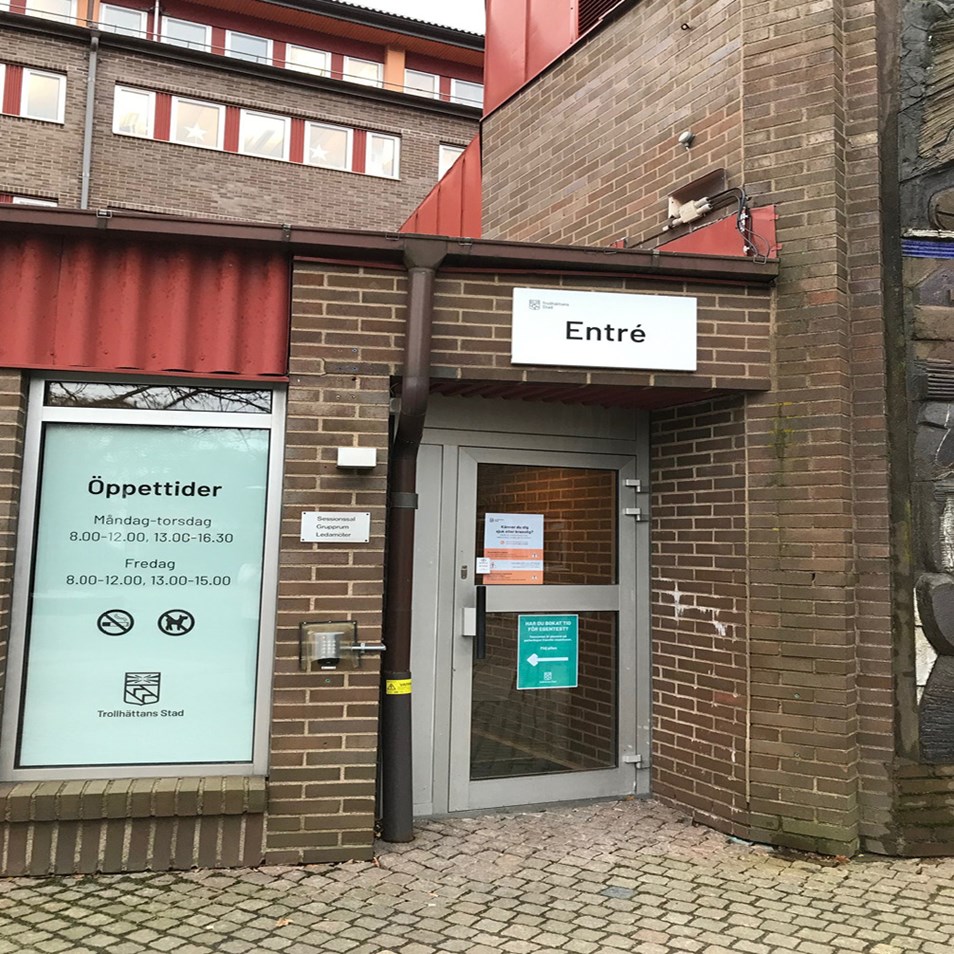 Bilden föreställer en mindre entré till Stadshuset. Dörren, som är i glas, är centrerad i bild. På dörren sitter två skyltar, en röd och en grön. Ovanför dörren sitter en stor skylt med texten entré. Till vänster om dörren sitter en jättestor skylt i fönstret som beskriver öppettiderna. 