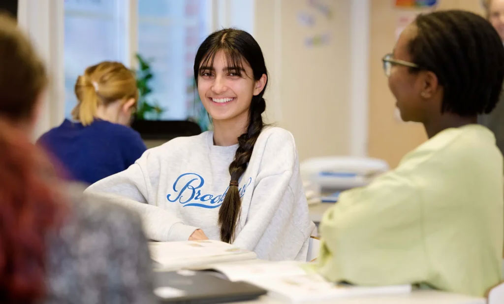 En tjej vid en skolbänk, skrattar och tittar in i kameran. Foto.