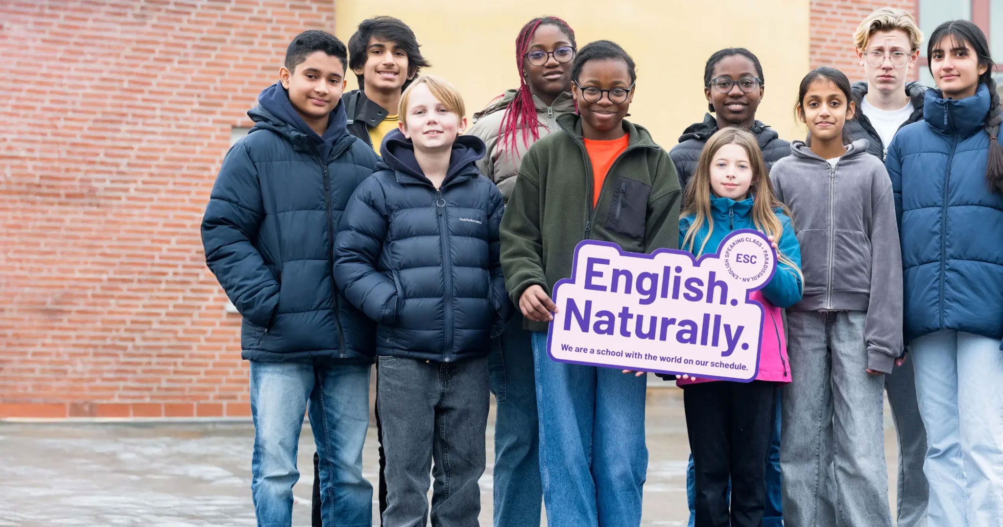 Tio skolelever står utomhus och håller upp en skylt med texten "English Naturally, we are a school with the world on our schedule.". Foto.