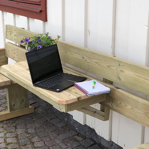 Fotografiet är en närbild av en svart laptop som står på ett bord som sitter fast i en vit vägg. Förutom datorn så står det en lila blomma på bordet och det ligger ett anteckningsblock och en penna bredvid. 