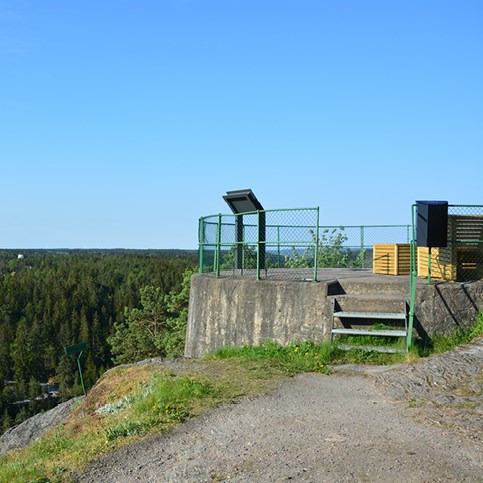 Bilden föreställer utsiktsplatsen Kopparklinten. Till höger i bild är en trappa upp till en avsats där det finns bänkar och en informationsskylt. Runt avsatsen är det staket i grönt. 