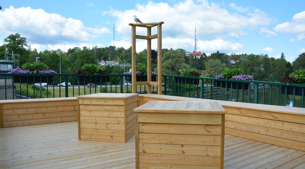 Bilden är ett fotografi av paviljongen vid Klaffbron. Paviljongen är en fyrkantig veranda med två bord i mitten. Paviljongen är byggd i trä. Det syns att den är ny då trädfärgen riktigt lyser. Det är sittplatser i form av bänkar runt hela paviljongen. 