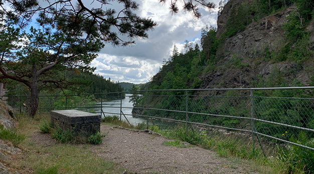 Bilden är ett fotografi över en sittplats utmed älven. Från platsen syns Göta älv och på båda sidorna älven är det berg och skog. 