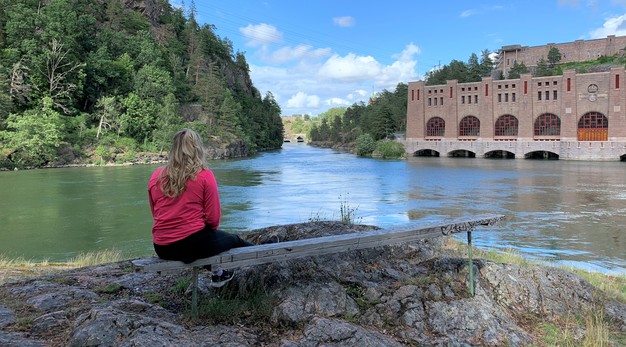 Bilden är ett fotografi på en kvinna som sitter på en bänk och tittar ut över Olidan. Vattnet är spegelblankt och det är soligt ute. Kvinna sitter längst ut på den bänkens vänstra sida. Hon är klädd i en rosa träningströja och svarta träningsbyxor. 