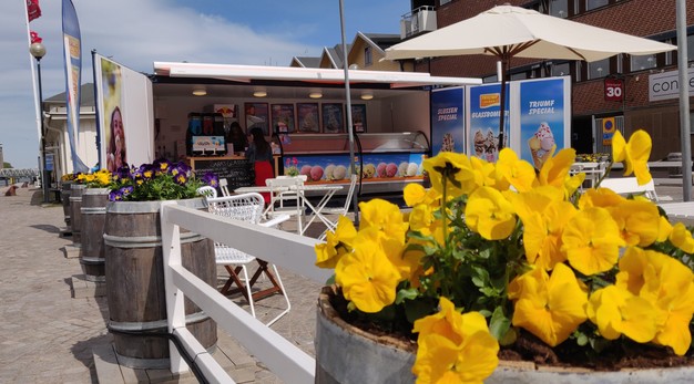 Blomplantering i förgrunden, cafébord och glasskiosk i bakgrunden. 