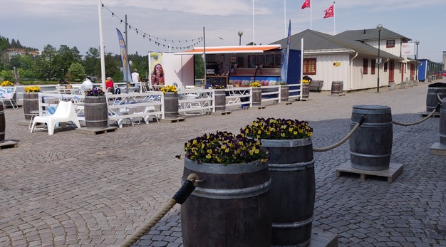 Bilden är ett fotografi över Strandgatans glasskiosk. I förgrunden syns två tunnor med gula och lila blommor. I bakgrunden syns klasskiosken till höger och uteserveringen till vänster. Runt kiosken och uteserveringen löper ett staket i vitt. 