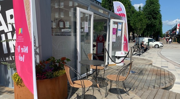 Bilden föreställer ingången till vårvikslokalen som blivit glasskiosk. Utanför lokalen finns en veranda formad som små cirklar. Utanför glasskiosken finns ett bord med tre stolar. 