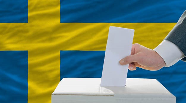 Hand som stoppar ner ett kuvert i en valurna. Svenska flaggan i bakgrunden