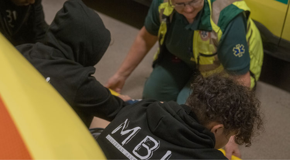 Bild på en ambulanssjuksköterska och två ryggtavlor på unga människor som lutar sig över en bår.