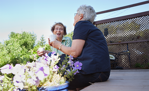 Två äldre kvinnor i trädgården dricker kaffe och skrattar tillsammans.