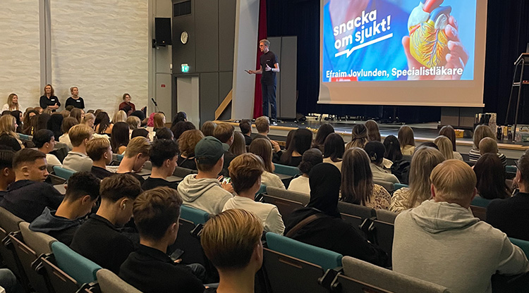 Bild på person som föreläser i en aula med massor av gymnasieelever i publiken