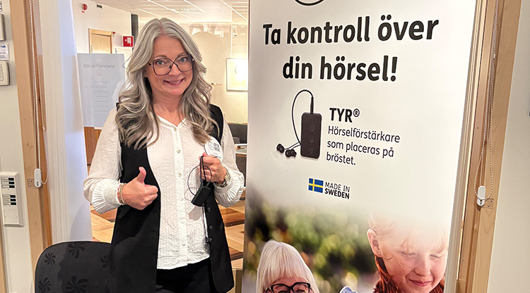 Kvinna ler och gör tummen upp till kameran. Står vid ett mässbord.
