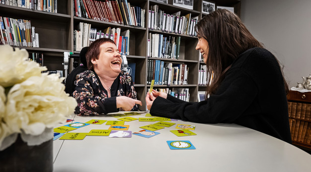 En personlig assistent sitter tillsammans med en person vid ett bord  i ett bibliotek och tittar på ett bildstöd tillsammans.