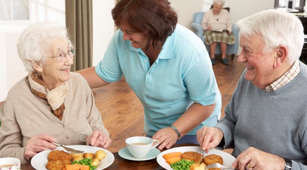 En äldre man och kvinna sitter och äter vid ett bord. En tredje person håller en hand på kvinnan och ler.