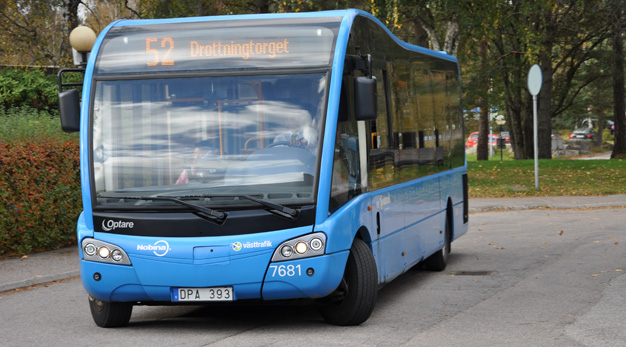 En blå buss åker på en gata. På sidan av gatan står några buskar.