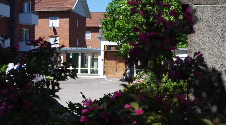 Stor gräsmatta med ett stort tegelhus i bakgrunden med balkonger. Blå himmel syns med några träd utspridda i balkongen
