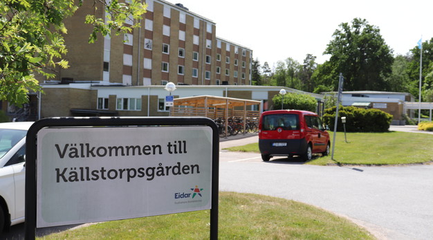 En skylt där det står välkommen till Källstorpsgården. Skylten står intill en parkering där två bilar syns. Bakom parkeringen ligger Källstorpsgården, ett stort flervåningshus i tegel.