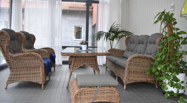 En soffa och ett bord står uppställt i en inglasad balkong. Mittemot soffan står två fåtöljer. I den inglasade balkongen finns även en pall samt en palm och en annan stor grön växt.