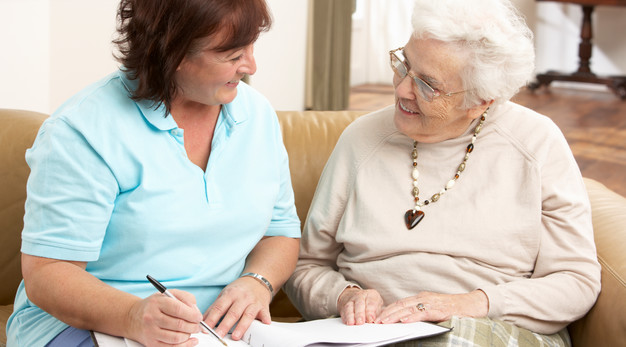 Äldre kvinna sitter i soffa i sitt hem tillsammans med vårdpersonal och samtalar glatt.