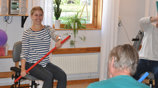 Två äldre män håller i varsin stav under en övning. Framför dem sitter en kvinnlig instruktör på en stol där hon håller i en egen stav.