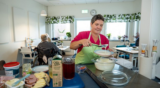 Vårdpersonal står i ett kök och förbereder mat. I bakgrunden sitter äldre personer vid bord.