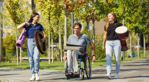Tre glada personer ute på promenad på väg till en aktivitet. En person sitter i rullstol.