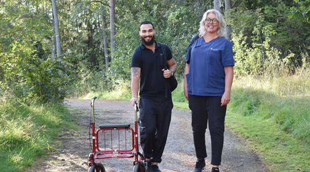 En Fysioterapeut och arbetsterapeut står på en cykelväg intill buskar och träd. Bredvid dem har de en rullator med sig.