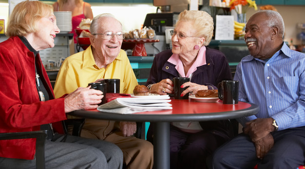 Fyra äldre personer sitter och dricker kaffe och fikar vid ett bord.