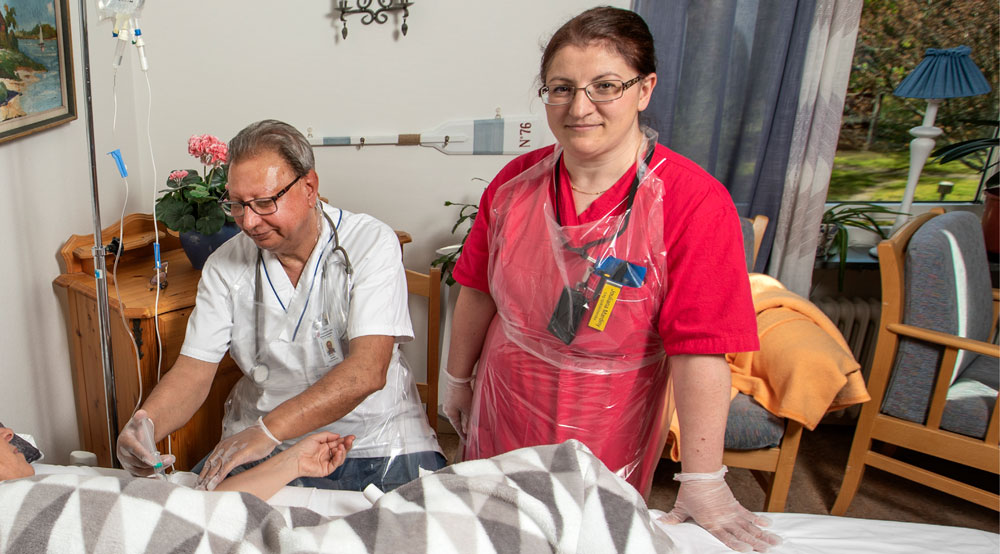 En sjuksköterska hjälper en patient i en säng med ett dropp. Bredvid står ytterligare en sjuksköterska.