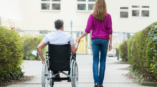 Kvinna promenerar utomhus tillsammans med en man i rullstol. De håller i varandras händer