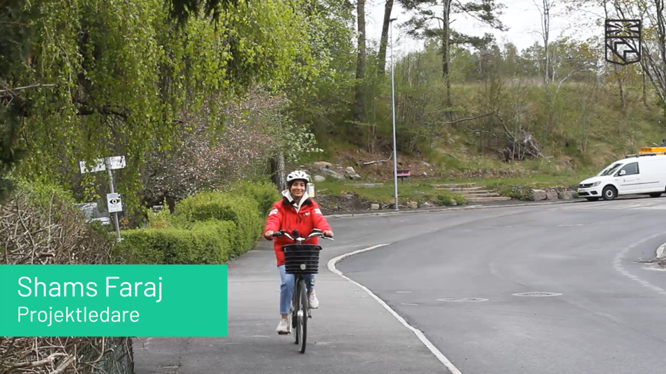 Projektledare Shams kommer cyklande i röd jacka och vit hjälm på en cykelväg. 