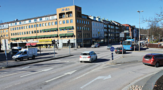 Fotot visaren vägkorsning med personbilar och en buss. En gul tegelbyggnad syns också i bakgrunden.