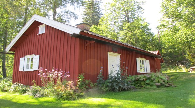Bilden föreställer en rött hus med vita knutar. Runt om huset växer blommor av olika sorter i lila och rosa. 