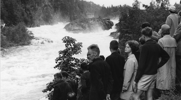 Besökare i 60-talskläder tittar ut över ett fallpåsläpp. Bilden är svartvit. 