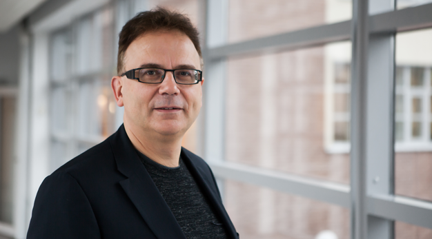 Porträtt på Martin Hellström, en mörkhårig man med glasögon. Han står inomhus med stora fönster i bakgrunden