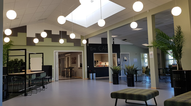 Ett rymligt rum med klotformade lampor i taket, stora växter och olika sittmöbler samt en servering/kiosk med skylten café.
