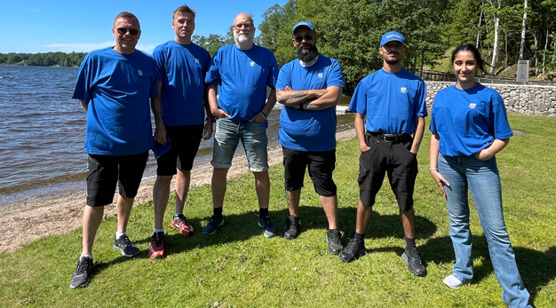 Sex personer står vid vattenkanten till en sjö iklädda likadana t-shirts med Trollhättans stads logga.