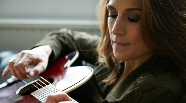 En stämningsfull bild där en mörkhårig Jill Johnson i grön jacka spelar på en gitarr.