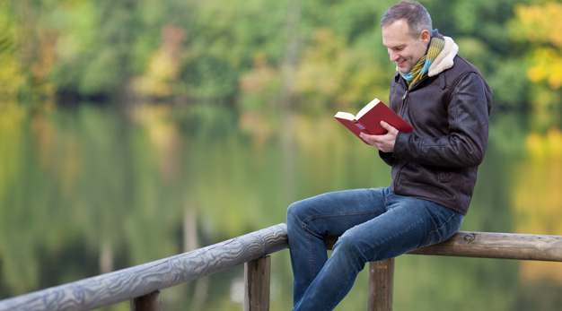 En man sitter på ett staket och läser bok. 