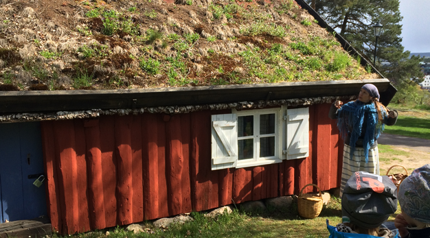 Pedagog iklädd gammaldags kläder visar ett litet rött hus med gräsbeklätt tak för små barn med mössa.