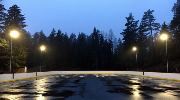 Asfalterad isrink med sarg och belysning. Skog syns i bakgrunden.