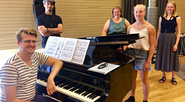 Fyra unga personer står runt en flygel. En manlig pianist sitter på pianostolen.