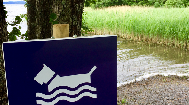 Bild på en skylt som visar hundbadplats med vatten i bakgrunden. 
