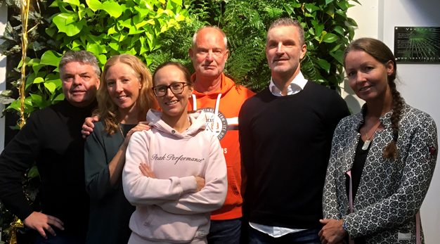 Gruppbild med tre män och tre kvinnor framför en vägg med växter.