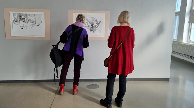 Två äldre kvinnor står och betraktar teckningar på en vägg. 