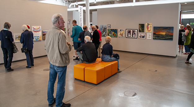 Många personer rör sig i utställningshallen och tittar på konstverken på väggarna. Några sitter och betraktar verken. 